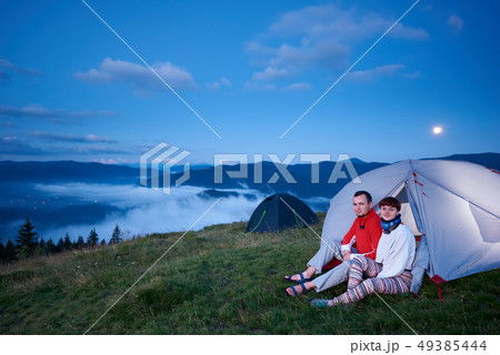 Guy and girl sitting in tent at dawn with view of beautiful landscape 49385444