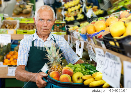 Senior male seller in apron putting fresh products on shelves in greengrocery Senior male seller in apron putting fresh products on shelves in greengrocery 49402164