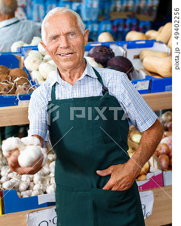 Positive senior male owner of greengrocery shop in apron offering fresh fruits and vegetables 49402256