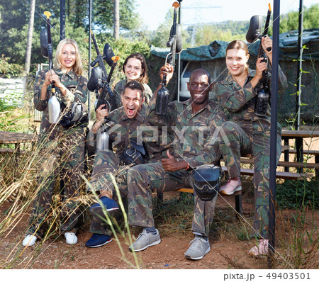 Friendly group of paintball players in camouflage standing with guns on paintball playing field outdoors 49403501