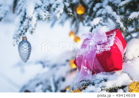 Closeup of a red gift box with a white ribbon next to Christmas decorations on the branches of a 49414639