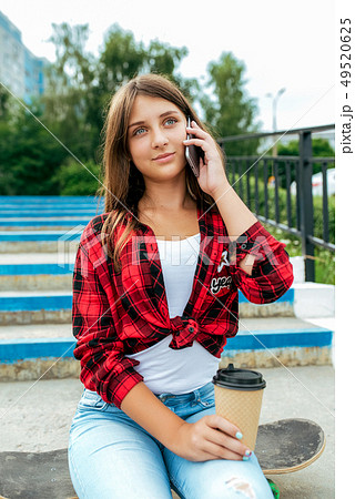 Girl teenager sits on a skateboard, talking on the phone, holding a cup with tea in her hands. In 49520625