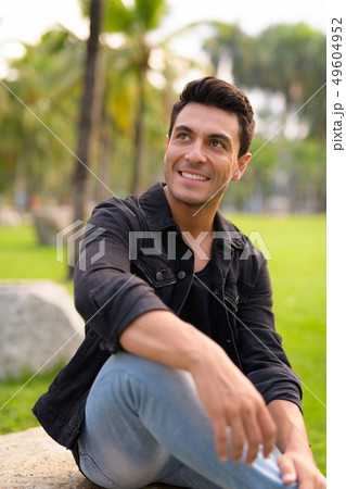 Happy young Hispanic man sitting and thinking at the park Happy young Hispanic man sitting and thinking at the park 49604952