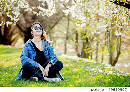 Young girl in a denim jacket and sunglasses sitting near a flowering tree Young girl in a denim jacket and sunglasses sitting near a flowering tree 49610997
