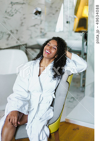 Brown skin relaxed girl with curly hair sitting in bathroom and wearing white bathrobe. 49634088