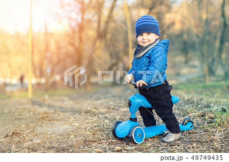 Cute adorable caucasian toddler boy in blue jacket having fun riding three-wheeled balance run bike Cute adorable caucasian toddler boy in blue jacket having fun riding three-wheeled balance run bike 49749435
