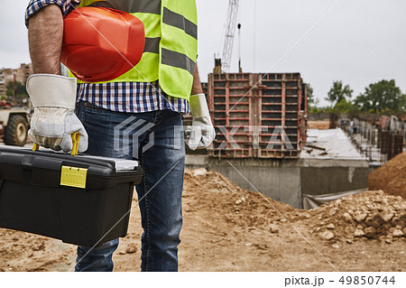 Ready to work! Cropped image of a builder in working uniform holding red helmet and carrying toolbox Ready to work! Cropped image of a builder in working uniform holding red helmet and carrying toolbox 49850744