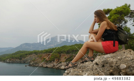Female tourist with backpack sitting on a rock on clear sky and calm sea background 49883382