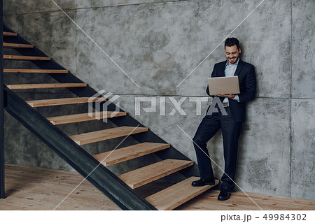 Smiling businessman is using laptop at staircase indoors Smiling businessman is using laptop at staircase indoors 49984302