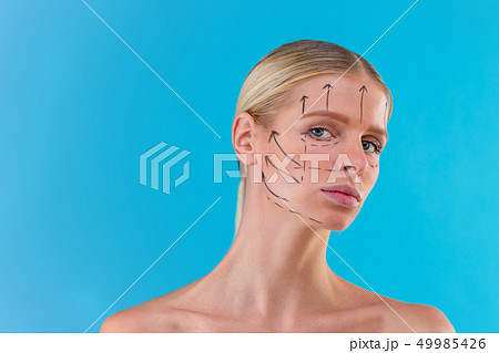 Beautician draw correction lines on woman face. Before plastic surgery operetion. Isolated on blue Beautician draw correction lines on woman face. Before plastic surgery operetion. Isolated on blue 49985426