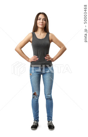 Front view of young attractive woman in gray sleeveless top and blue jeans standing with arms on 50003468