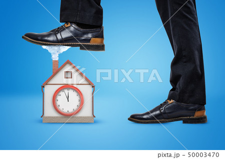 Side crop close-up of man's legs, one foot raised to step on a little house with a big clock on the Side crop close-up of man's legs, one foot raised to step on a little house with a big clock on the 50003470