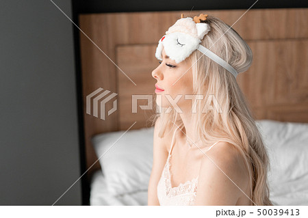 Morning in hotel room. Young woman sits on comfortable bed in mask for sleeping on head. White Morning in hotel room. Young woman sits on comfortable bed in mask for sleeping on head. White 50039413