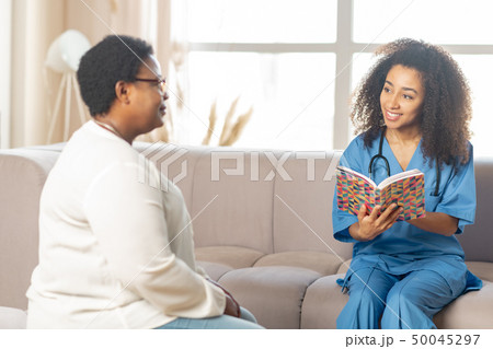 Pleasant dark-eyed sick nurse reading book for sick aged woman Pleasant dark-eyed sick nurse reading book for sick aged woman 50045297