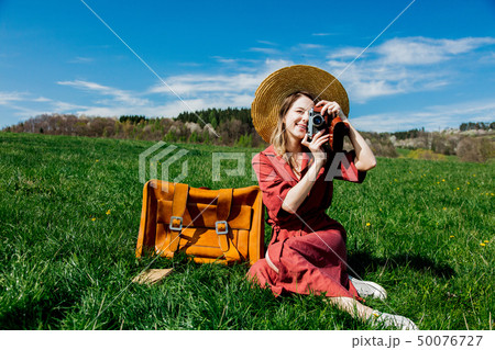 Beautiful girl in red dress and hat sitting on meadow with vintage camera and suitcase. 50076727