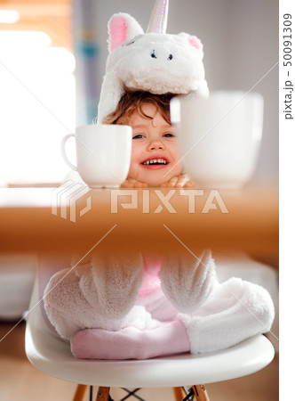 A portrait of small girl in unicorn mask sitting at the table at home. 50091309