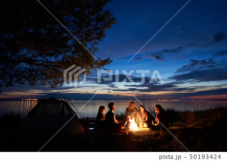 Night summer camping on shore. Group of young tourists around campfire near tent under evening sky 50193424