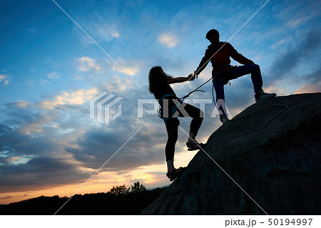 woman with rope engaged in sports of rock climbing on the top of cliff. Her friend helping her 50194997