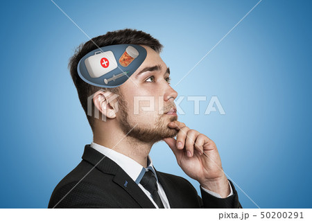Young businessman thinking with first aid medical box, pills jar and syringe in his head on blue Young businessman thinking with first aid medical box, pills jar and syringe in his head on blue 50200291