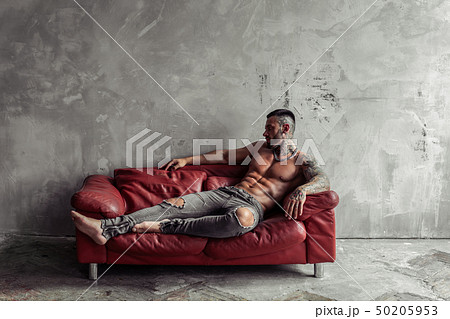 Fashion portrait of Sexy naked male model with tattoo and a black beard lying in hot pose on red Fashion portrait of Sexy naked male model with tattoo and a black beard lying in hot pose on red 50205953