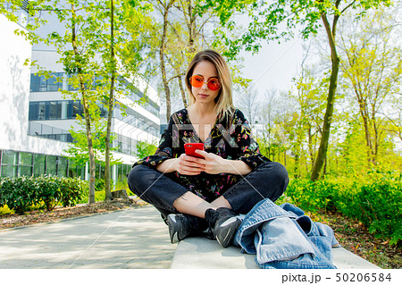 Young woman in sunglasses with mobile phone sitting on green alley near modern building of Young woman in sunglasses with mobile phone sitting on green alley near modern building of 50206584