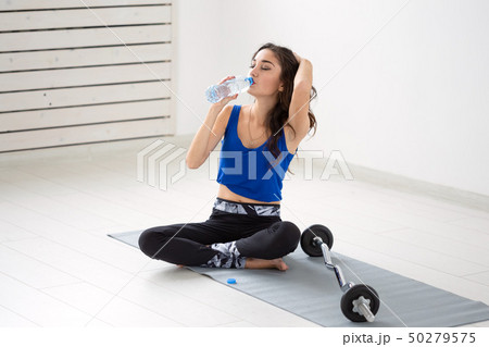 Healthy lifestyle, people and sport concept - Smiling woman sitting on yoga mat and drinking water Healthy lifestyle, people and sport concept - Smiling woman sitting on yoga mat and drinking water 50279575