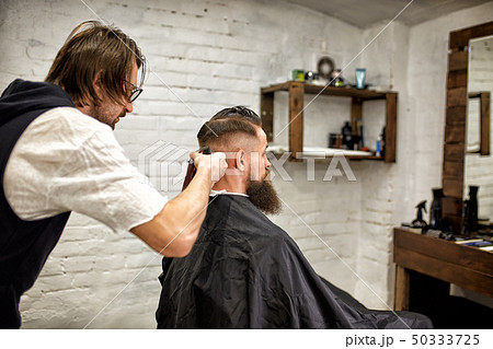 Brutal guy in modern Barber Shop. Hairdresser makes hairstyle a man with a long beard. Master Brutal guy in modern Barber Shop. Hairdresser makes hairstyle a man with a long beard. Master 50333725