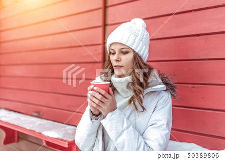 Beautiful girl in winter in city on background of red shutters. In his hand he holds a glass with a Beautiful girl in winter in city on background of red shutters. In his hand he holds a glass with a 50389806