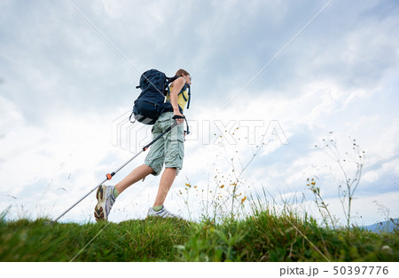 Woman hiker hiking on grassy hill, wearing backpack, using trekking sticks in the mountains 50397776