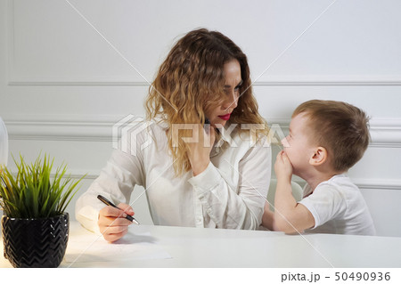 Little boy is stopping his mom to work while she is calling phone in her workplace in office 50490936