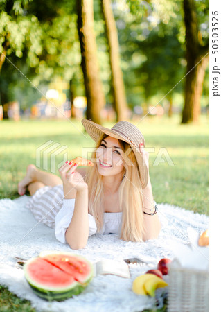 Young smiling girl in hat with croissant lying in park on plaid near watermelon. 50503526