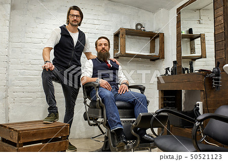 Barbershop, hairdresser and client. Hairdresser and bearded young guy posing for the camera before a 50521123