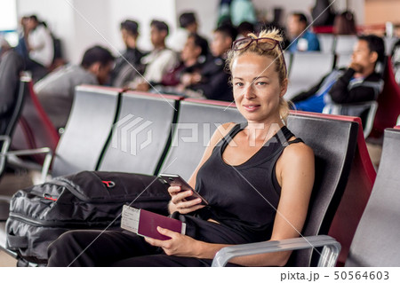 Casual tanned blond female traveler holding cell phone, passport and boarding pass while waiting to 50564603
