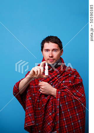 A wrapped in a blanket sick man is standing with nasal spray in his hand 50590385