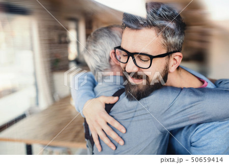 An adult hipster son and senior father indoors at home, hugging. 50659414