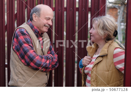 Two senior neighbors takling to each other on sunny day near fence. Two senior neighbors takling to each other on sunny day near fence. 50677128