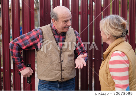 Two senior neighbors takling to each other on sunny day near fence. Two senior neighbors takling to each other on sunny day near fence. 50678099