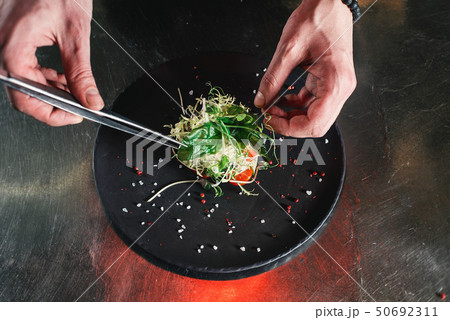 Chef finishing healthy salad on a black plate with tweezers. almost ready to serve it on a table 50692311