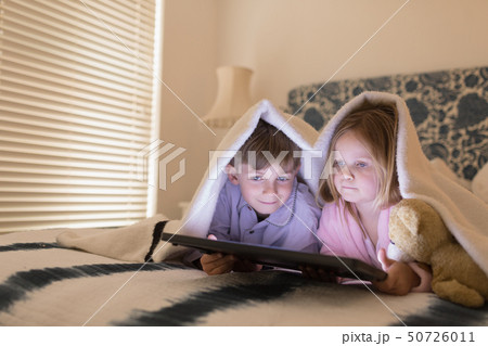 Siblings using digital tablet under blanket in bedroom Siblings using digital tablet under blanket in bedroom 50726011
