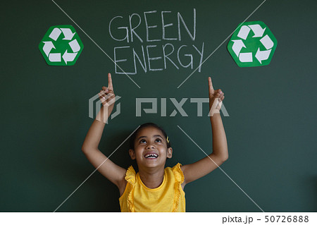 Smiling schoolgirl with arms up standing against green energy board in classroom 50726888