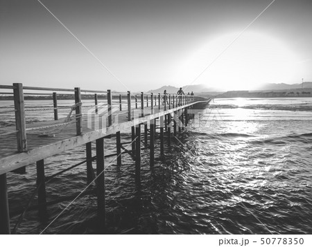 Black and white image of sun setting down over the ocean waves and long wooden pier Black and white image of sun setting down over the ocean waves and long wooden pier 50778350