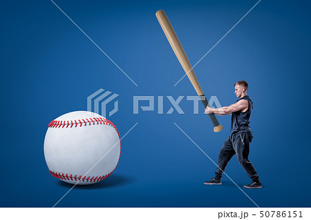 Side view of young athletic man in gym suit, holding enormous bat and ready to hit huge baseball. 50786151