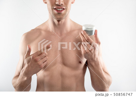 Young man with perfect body holding jar of cosmetic cream and pointing at it Young man with perfect body holding jar of cosmetic cream and pointing at it 50856747