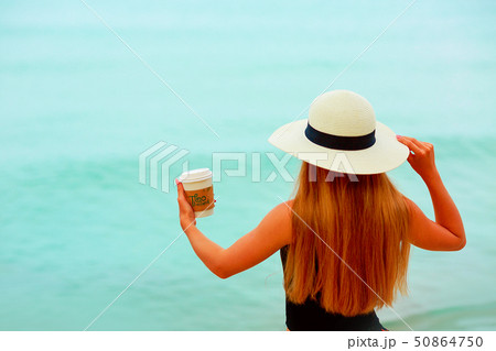 Woman in hat with a plastick glass of coffee is standing on the beach 50864750