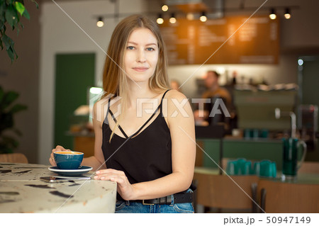 Young beautiful blonde with long hair in a charm top and jeans drinks coffee and looks out the 50947149