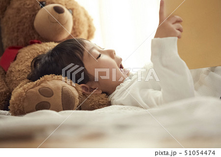 ベッドで大きなクマを枕に絵本を読む女の子 51054474