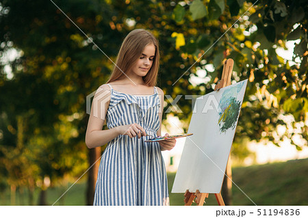 Beautiful girl artist is standing in the park in front of picture and holding palette with paints Beautiful girl artist is standing in the park in front of picture and holding palette with paints 51194836