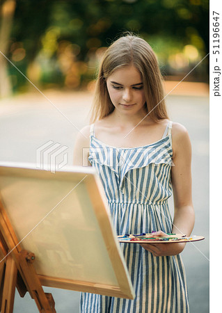 Beautiful girl draws a picture in the park using a palette with paints and a spatula. Easel and Beautiful girl draws a picture in the park using a palette with paints and a spatula. Easel and 51196647