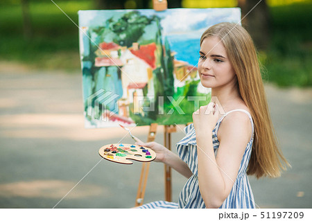 Beautiful blond hair girl sitting on stool and draws a picture in the park using a palette with Beautiful blond hair girl sitting on stool and draws a picture in the park using a palette with 51197209