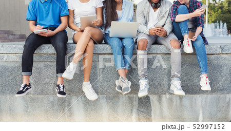 Company of teen friends sitting outdoor and using digital gadgets 52997152